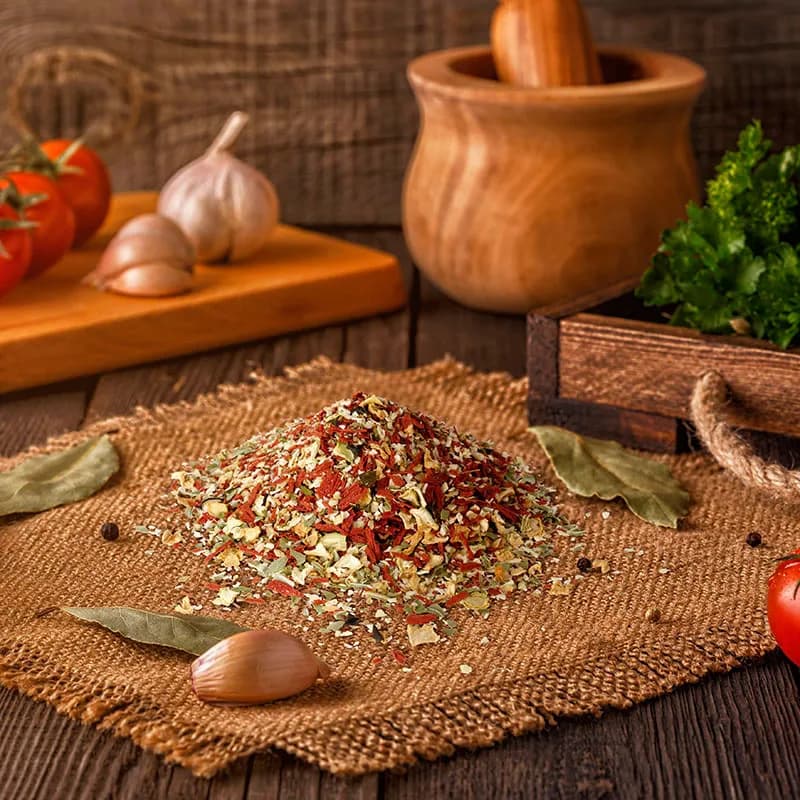 Gewürzmischung auf brauner Stoffmatte mit Mörser, Tomaten und Knoblauch, Lifestyle-Aufnahme