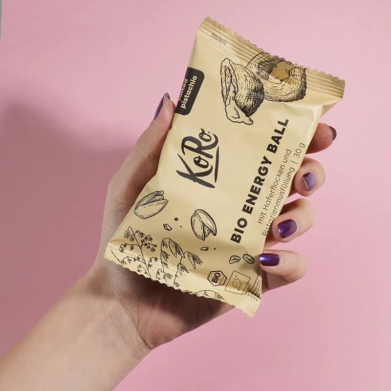 Hand hält eine Energy-Ball-Packung vor rosafarbenem Hintergrund, Mood-Aufnahme