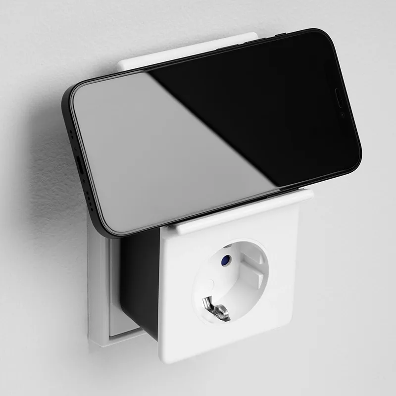 Smartphone liegt in einer weißen Ablage, die auf eine Wandsteckdose gesteckt ist, Mood-Aufnahme vor heller Wand