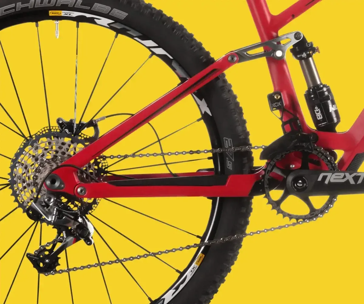 Nahaufnahme am Mountainbike: roter Hinterbau mit Kassette, Schaltwerk, Kette und Dämpfer vor gelbem Grund