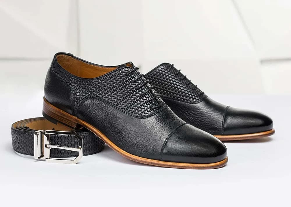Schwarze Oxford-Schuhe aus Leder mit Flechtmuster und passender Gürtel auf hellem Grund