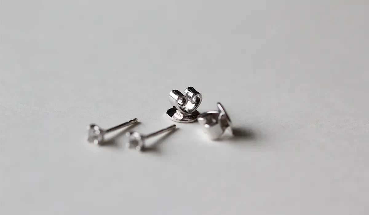 Nahaufnahme von zwei silbernen Ohrsteckern mit kleinen Diamanten und ihren passenden Verschlüssen auf einer weißen Oberfläche. Die geringe Tiefenschärfe betont die Vorderseite der Ohrringe.