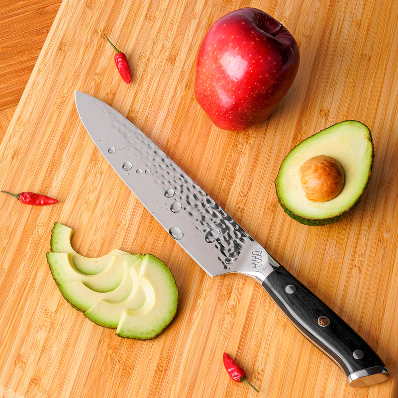 Professionelle Mood-Fotografie eines Messers mit schwarzem Griff auf einem Schneidebrett aus Holz dekoriert mit Apfel, Avocado und Chilis.
