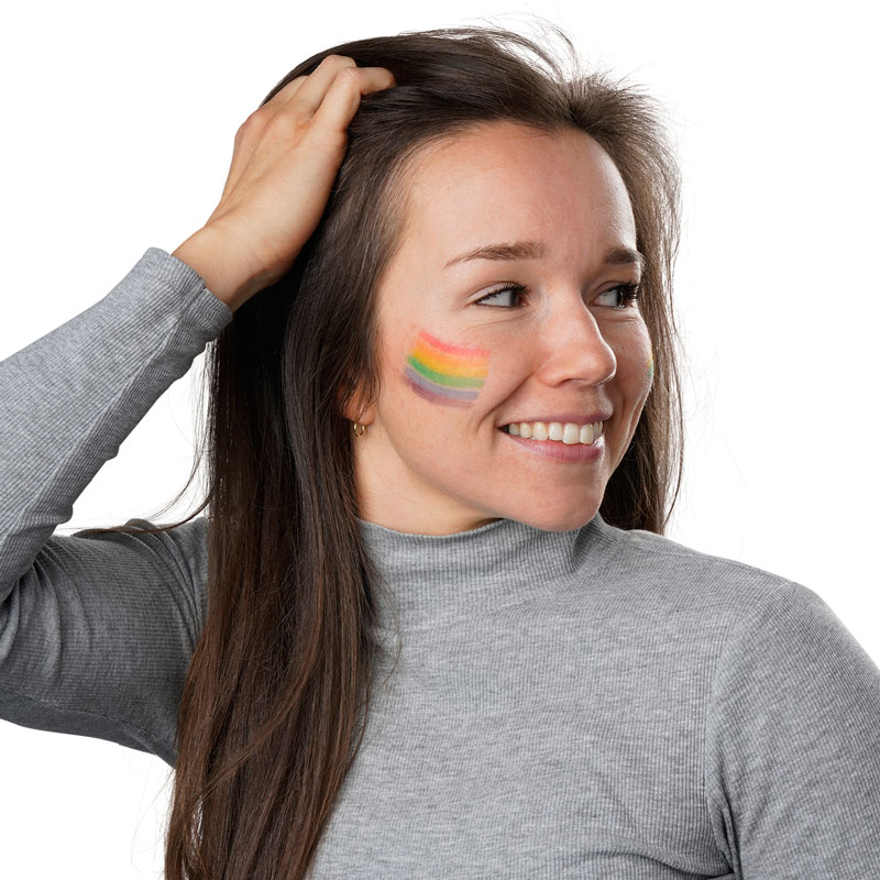 Professionelle Fotografie einer Frau, die sich mit Ihrer Hand durch die Haare fährt, einen grauen Rollkragen-Pullover und eine Regenbogenflagge auf der rechten Wange trägt.