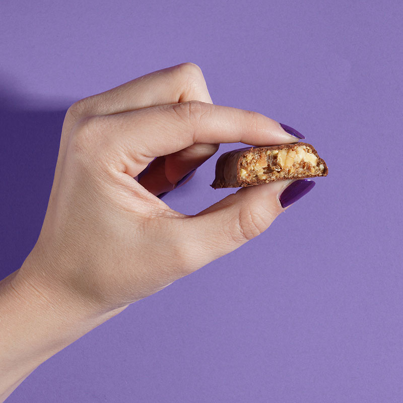 Professionelle Lifestyle-Fotografie eines Proteinriegels, welcher zwischen Daumen und Zeigefinger einer Frauenhand mit lackierten Nägeln vor lila Hintergrund gehalten wird.